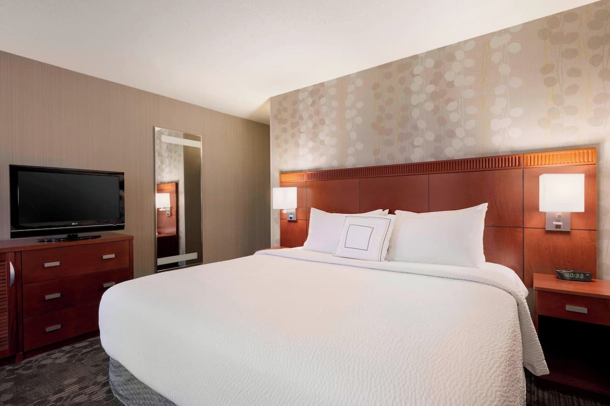 A hotel room with a large bed, a TV on a wooden dresser, and a nightstand with a clock.