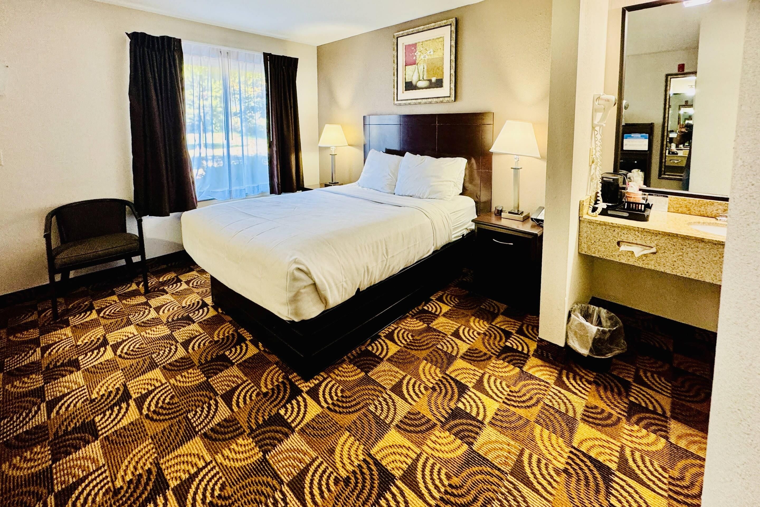 A hotel room with a bed, a chair, a desk with a lamp, a mirror, and a patterned carpet.