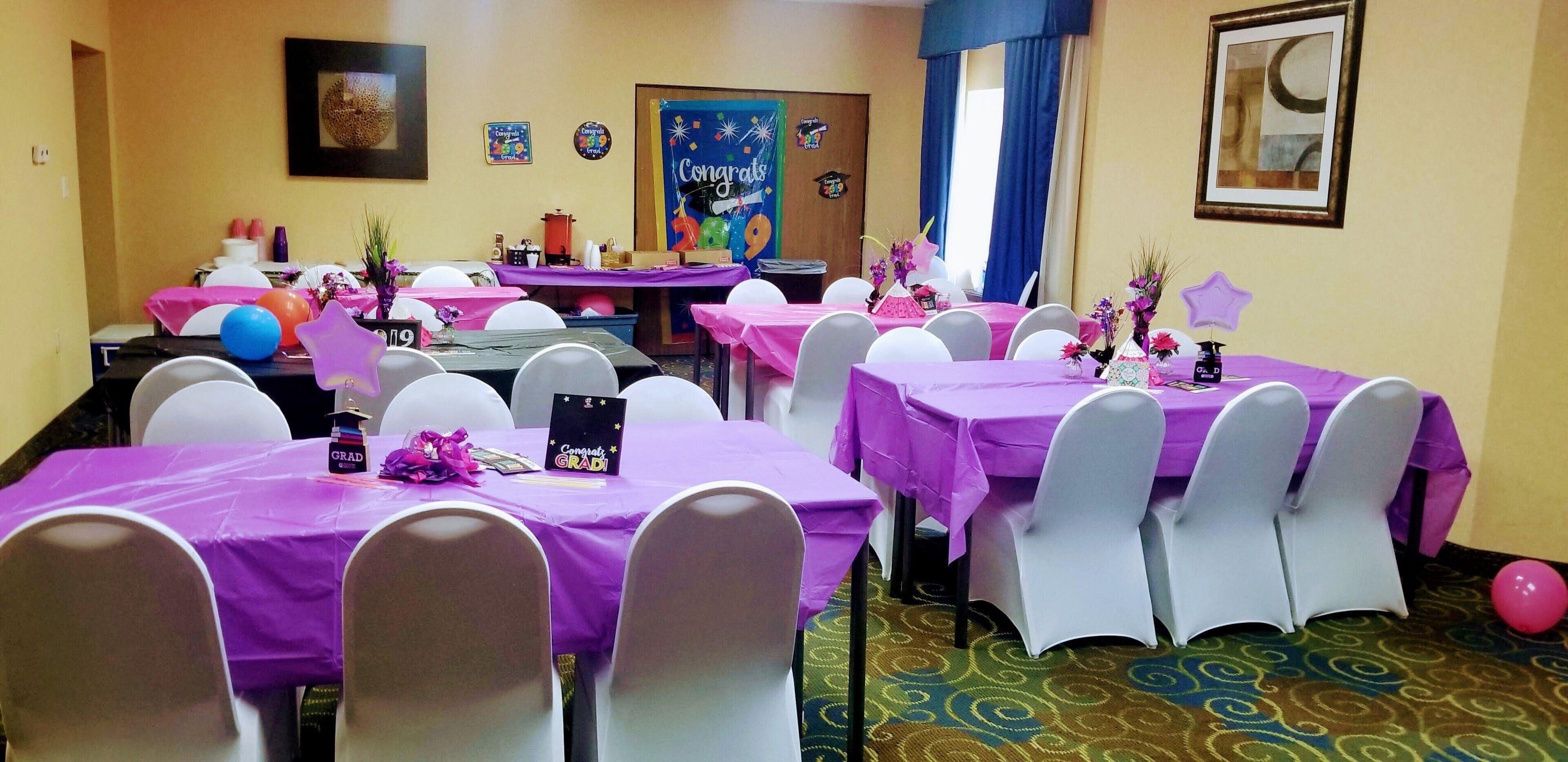 Una stanza con diversi tavoli ricoperti da tovaglie viola, sedie bianche e varie decorazioni, tra cui palloncini e uno striscione con la scritta 'Congratulations'.
