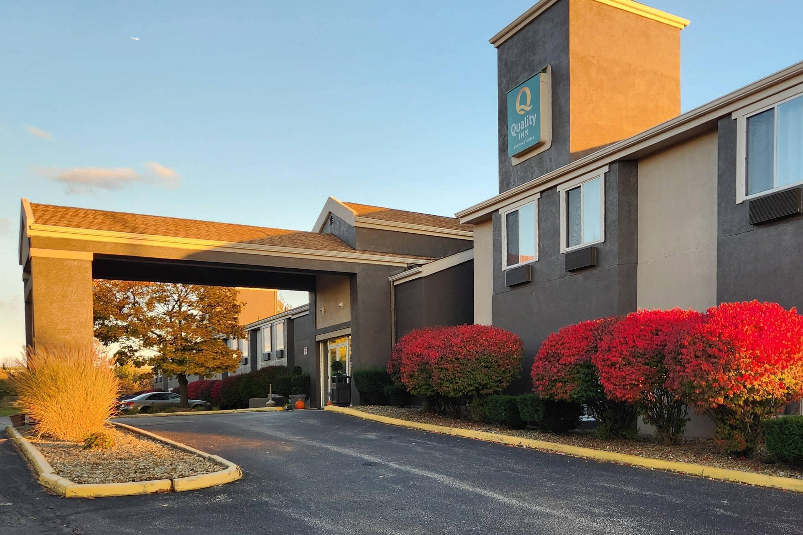 L’entrée d’un hôtel, avec une allée couverte, une enseigne indiquant « Quality Inn » et des arbustes à fleurs rouges.