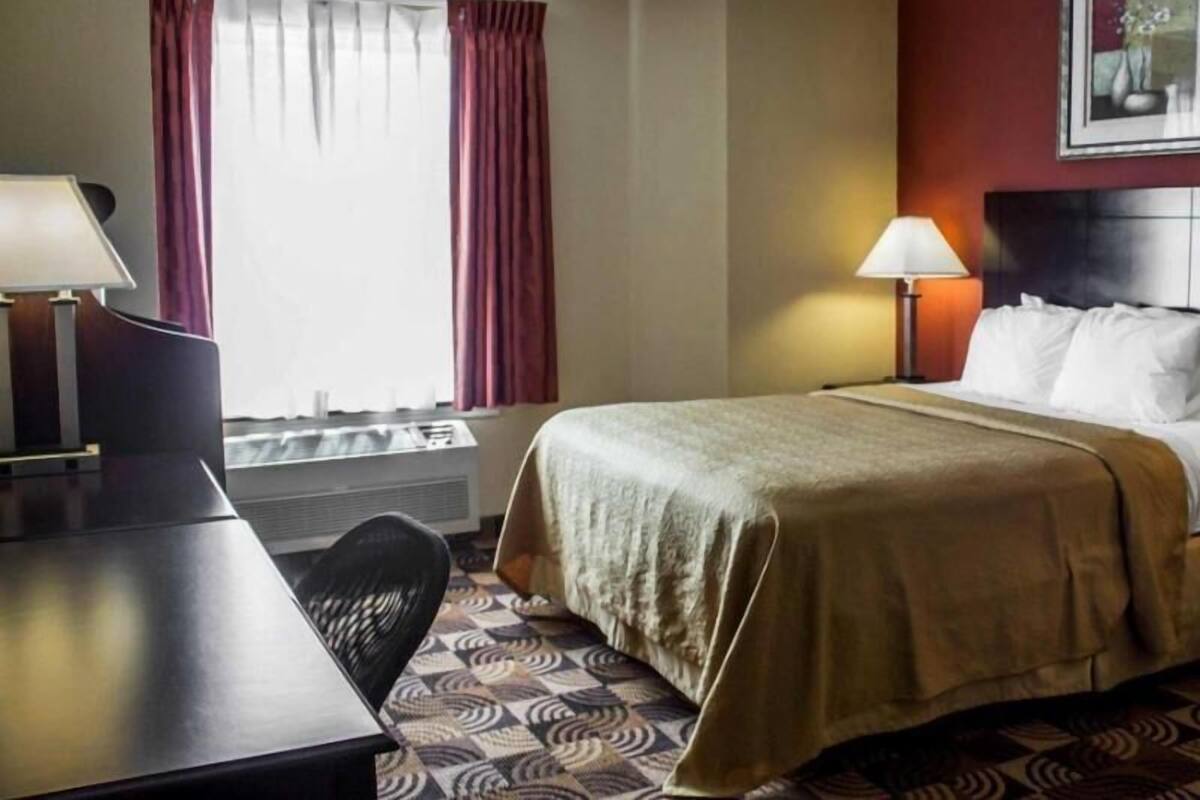 A hotel room with a bed, desk, chair, window with curtains, and a painting on the wall.