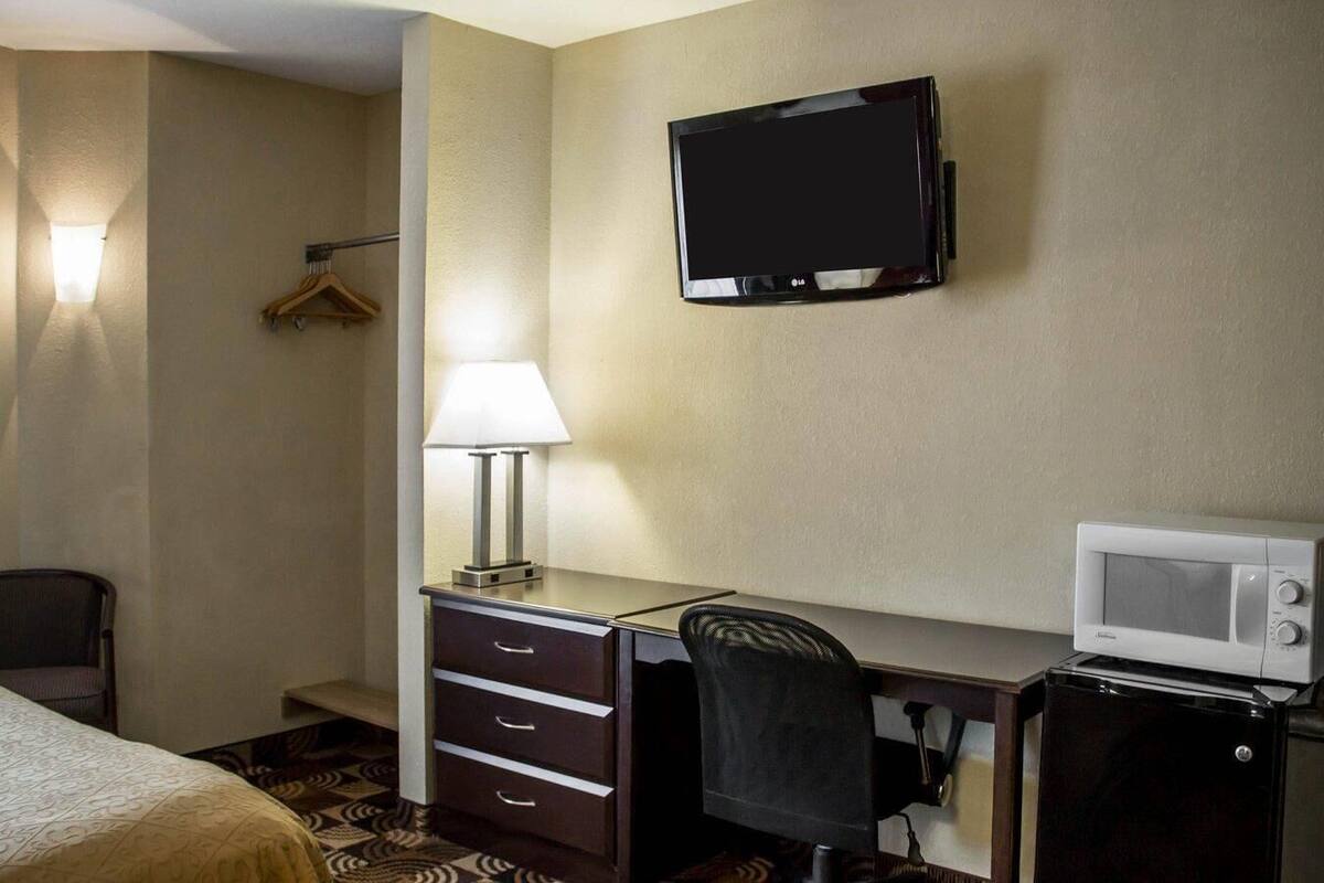 A hotel room with a desk, a chair, a TV, a lamp, a microwave, and a bed.