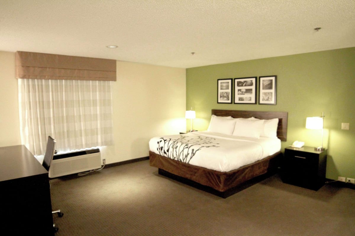 A hotel room with a bed, two bedside tables with lamps, a desk with a computer, and a window with blinds.