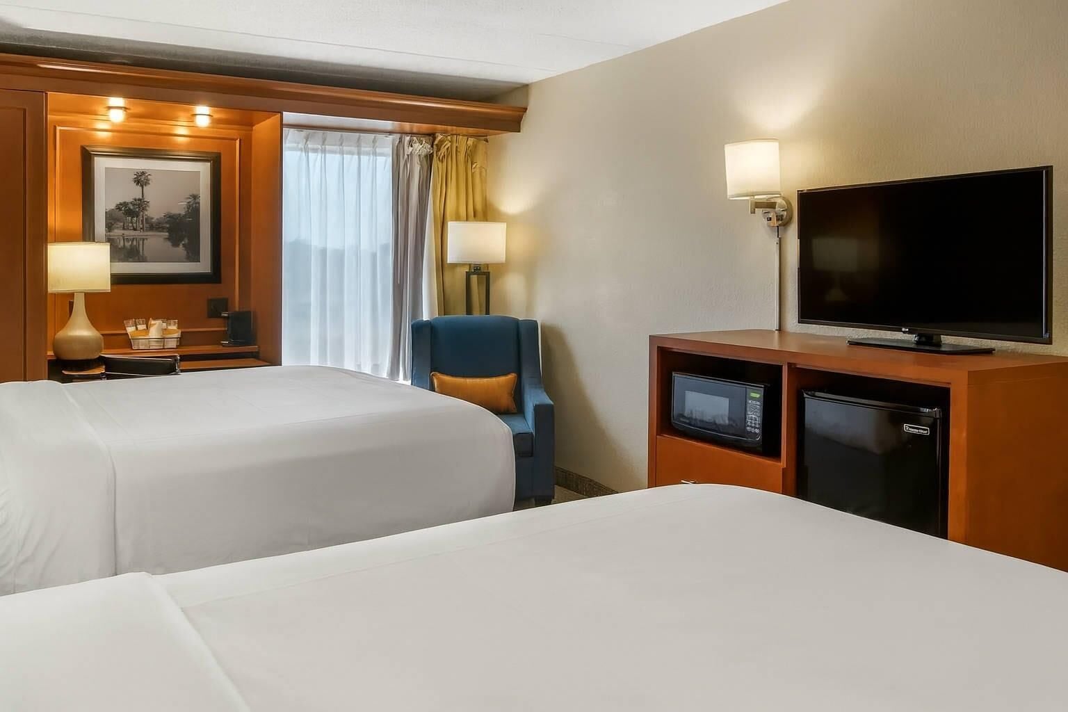 A hotel room with two beds, a desk with a lamp, a television, a mini-fridge, and a chair.