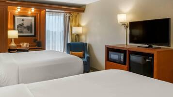 A hotel room with two beds, a desk with a lamp, a television, a mini-fridge, and a chair.