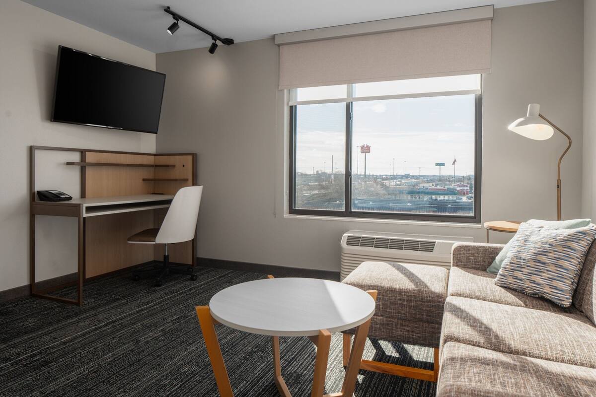 A modern hotel room with a desk, chair, and couch. A flat-screen TV is mounted on the wall, and a large window offers a view of the city.
