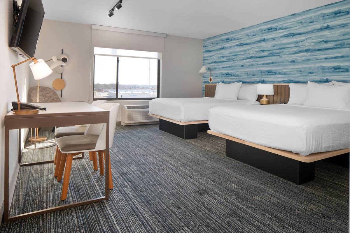 A hotel room with a desk, two beds, a large window, and a blue textured wall.