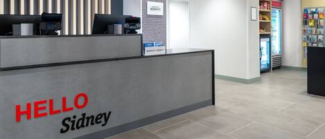 A reception desk with a 'HELLO Sidney' sign, a self-service kiosk, a minibar, and a vending machine.