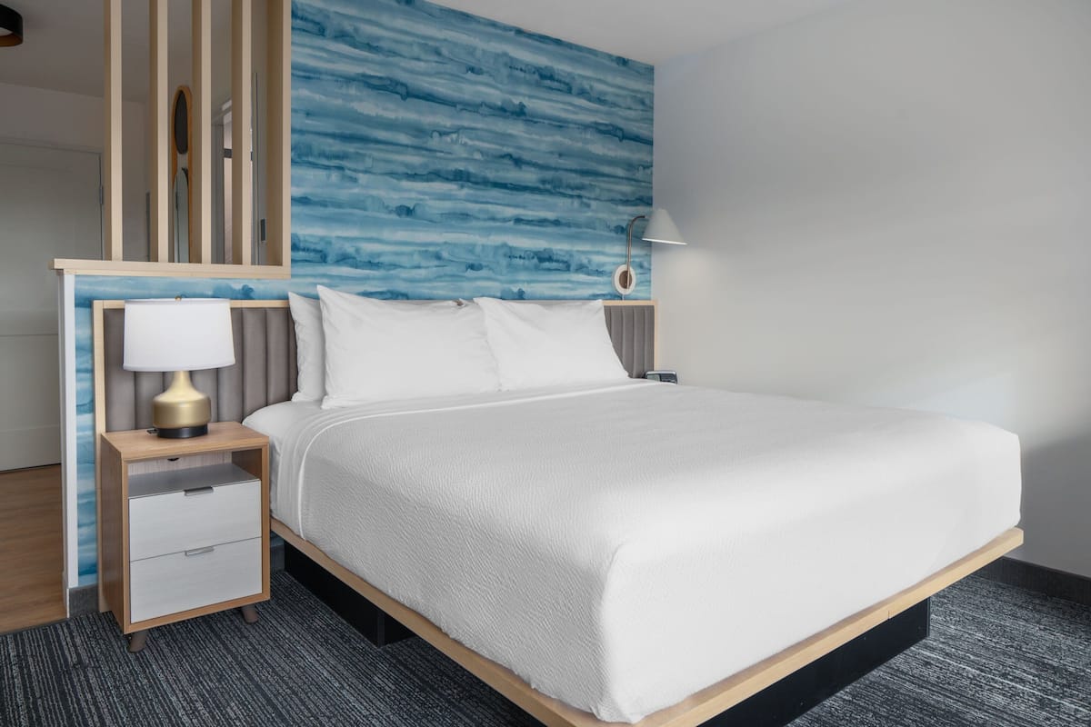 A neatly made bed with white linens, a wooden nightstand with a lamp, and a wall with a blue abstract pattern.