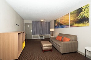 A hotel room with a grey sofa, an ottoman, a wooden desk, a wall-mounted air conditioner, and a large window with curtains. - Days Inn & Suites by Wyndham Dayton North (Dayton)