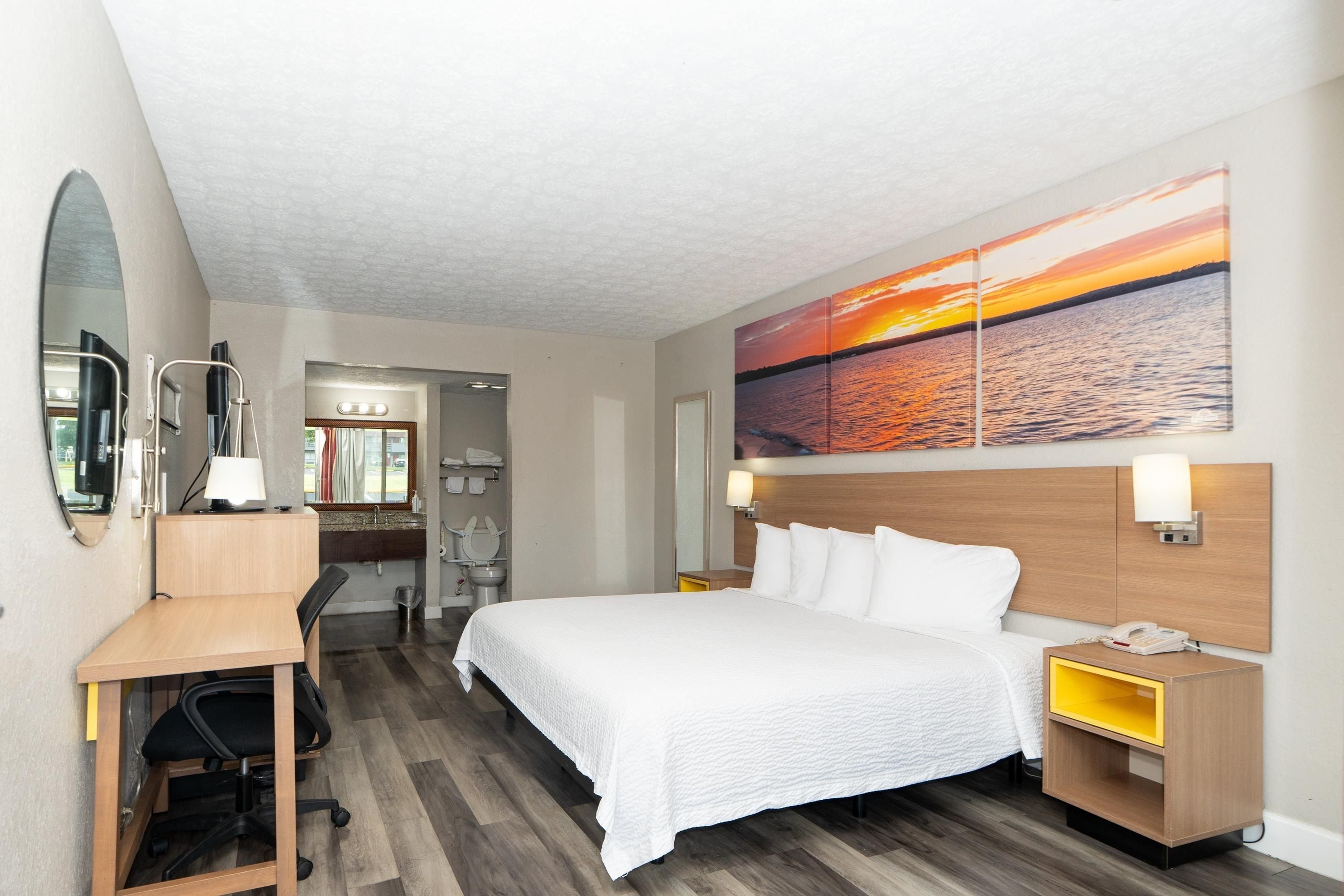 A hotel room with a bed, desk, chair, and a large painting of a sunset over water.