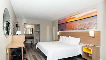 A hotel room with a bed, desk, chair, and a large painting of a sunset over water.
