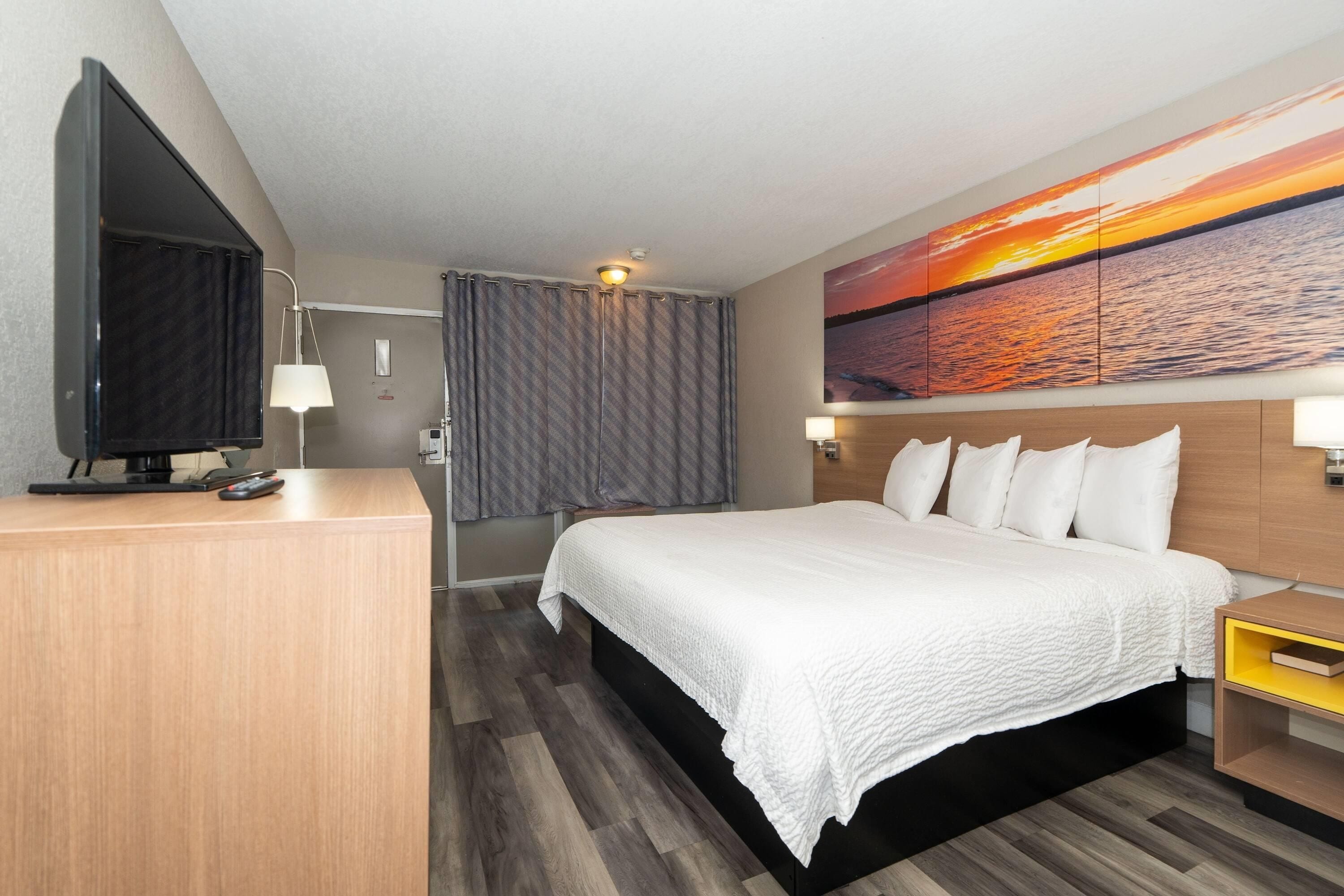 A hotel room with a bed, a TV, a desk, and a painting of a sunset.
