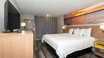 A hotel room with a bed, a TV, a desk, and a painting of a sunset.