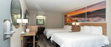 Habitación de hotel con dos camas, un escritorio, una silla, un cesto de basura, un espejo y un cuadro grande con un atardecer sobre el océano.