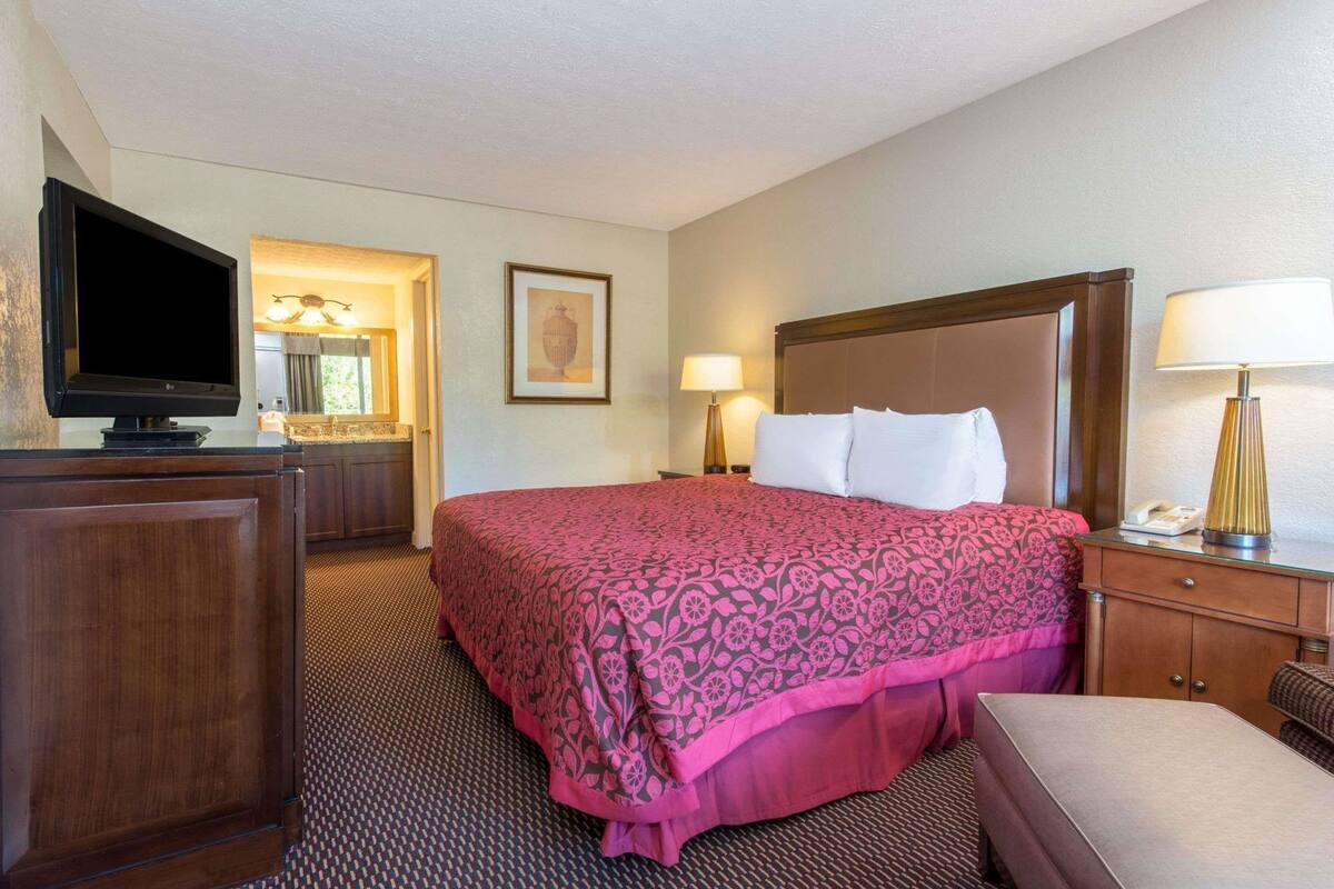 A hotel room with a bed, a TV, a desk, a lamp, and a painting on the wall.