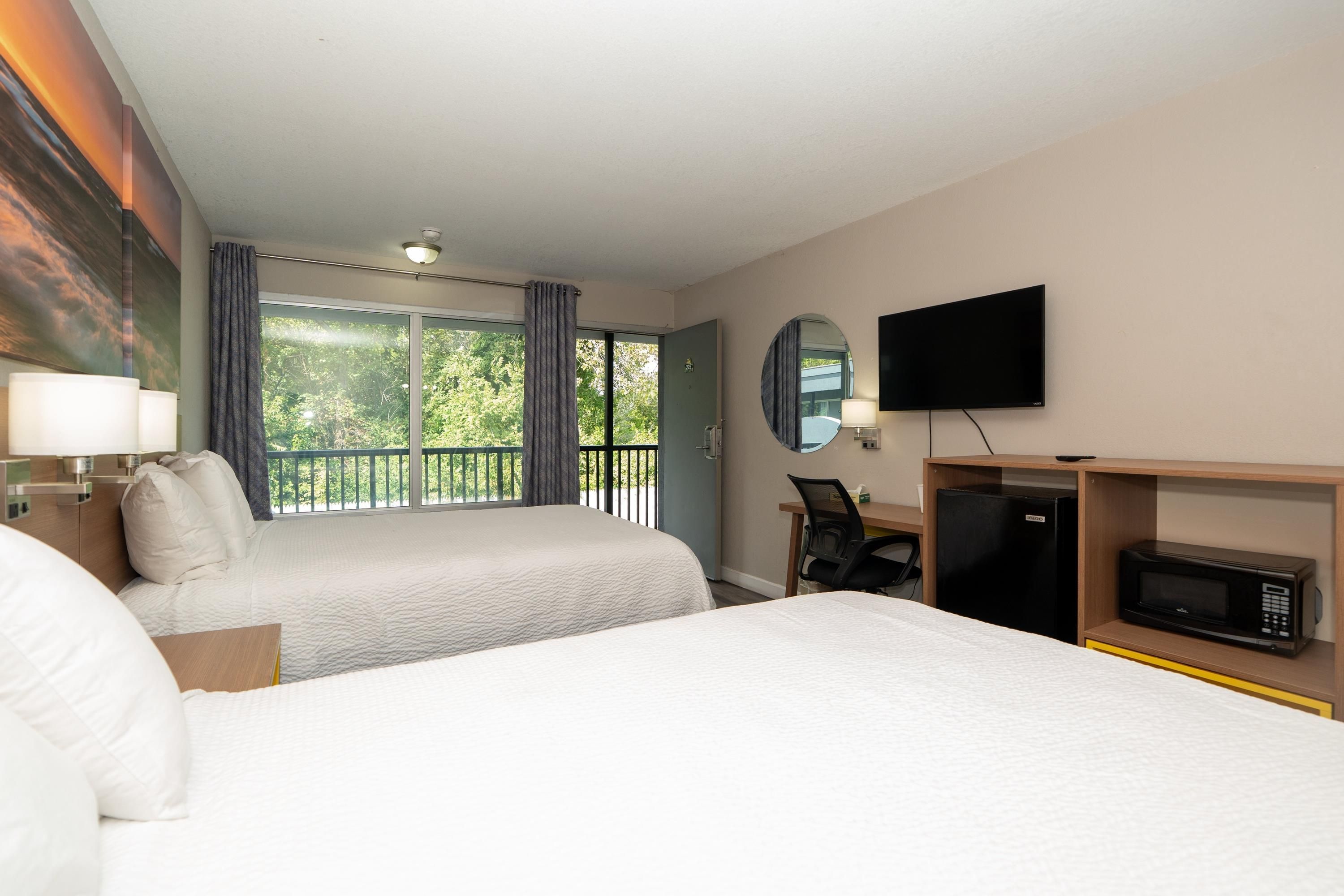 Quarto de hotel com duas camas, televisão, escrivaninha com cadeira, micro-ondas e varanda com cortinas.