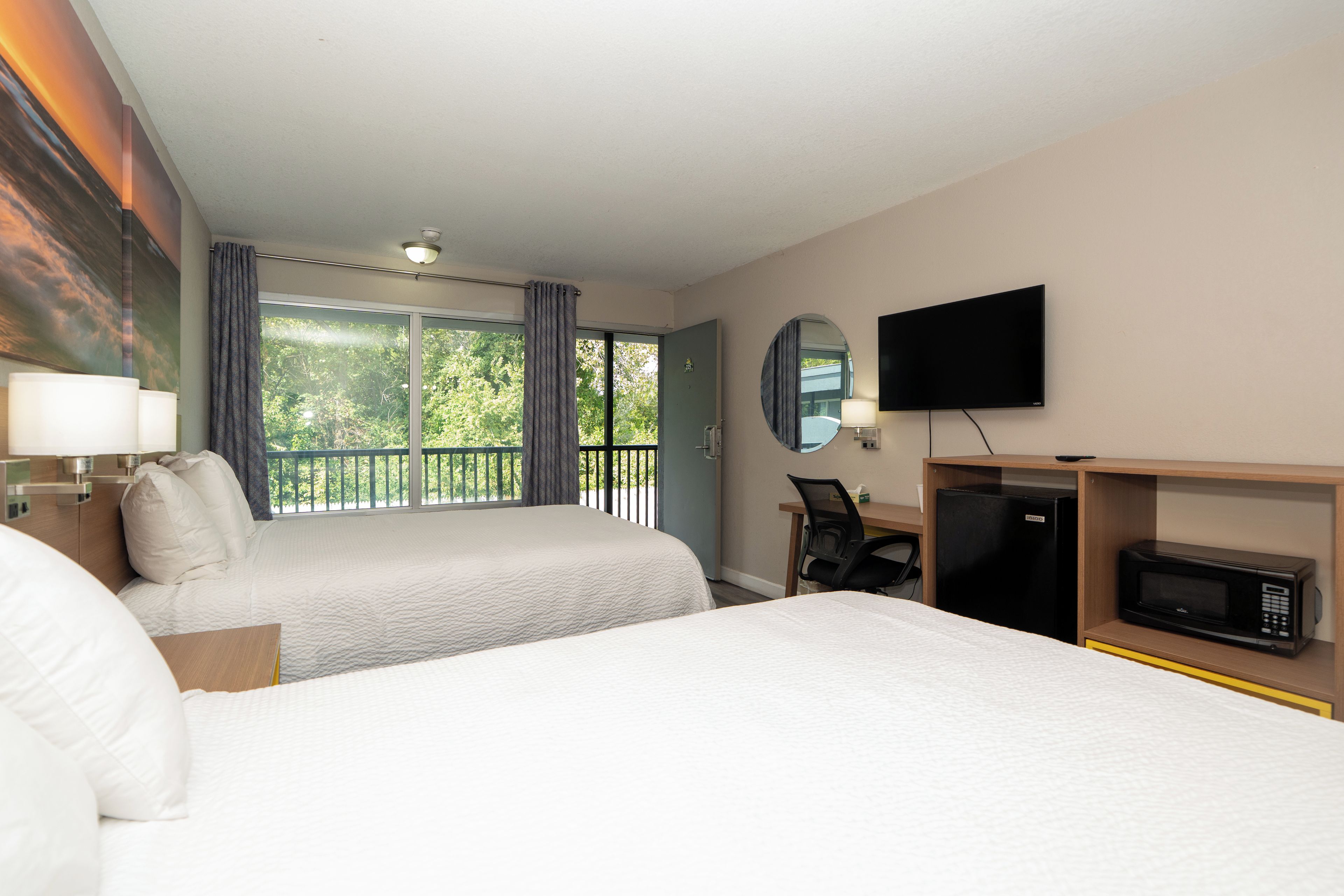 A hotel room with two beds, a desk with a chair, a television, a microwave, and a balcony with curtains.