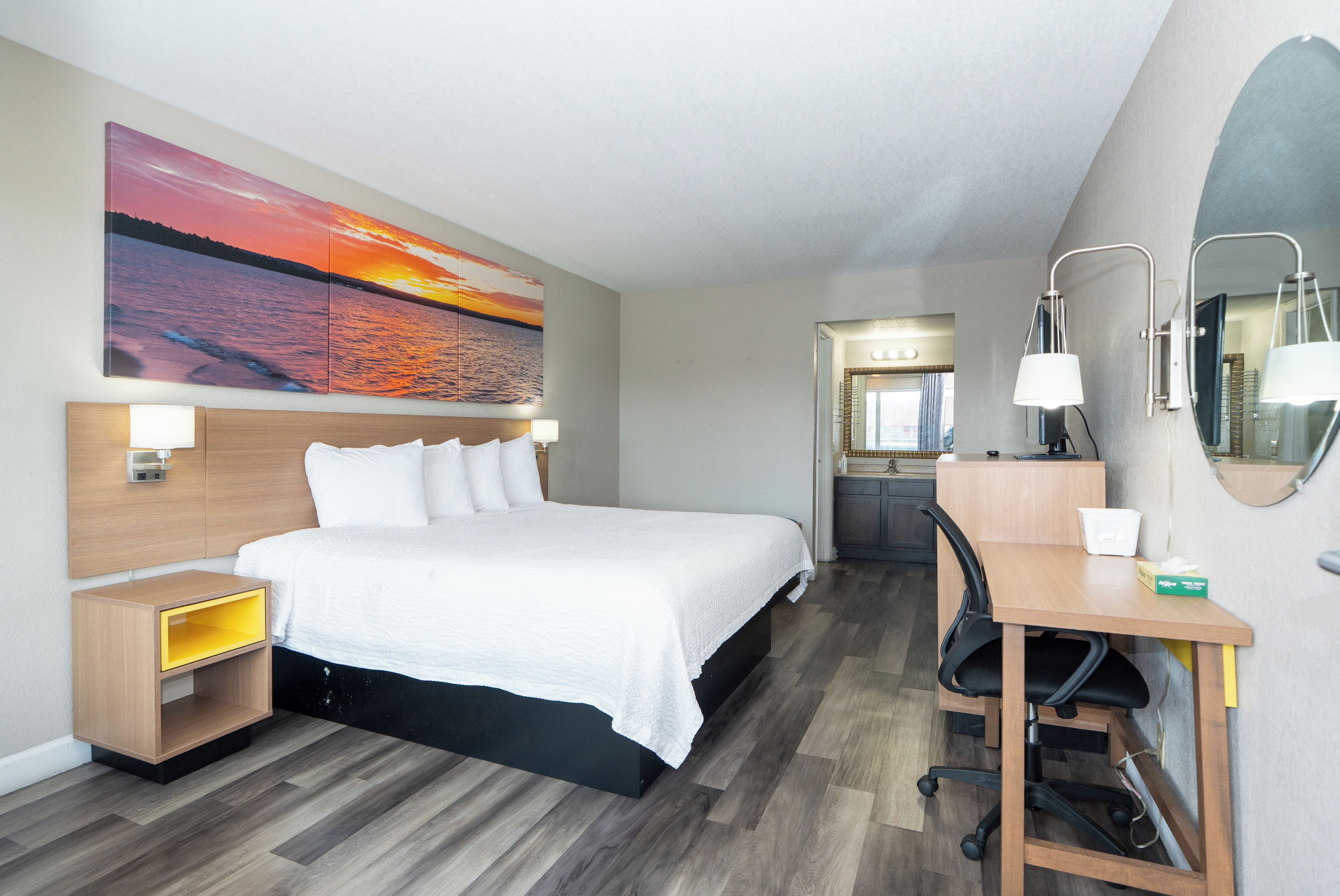 Habitación de hotel con cama, mesitas de noche, escritorio con silla, lámpara de escritorio, espejo y un cuadro grande con una puesta de sol sobre el agua.