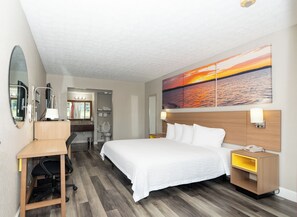 A hotel room with a bed, desk, chair, and a painting of a sunset. - Days Inn & Suites by Wyndham Dayton North (Dayton)
