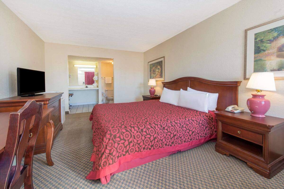 A hotel room with a bed, a desk with a television, a nightstand with a lamp, and a bathroom visible in the background.