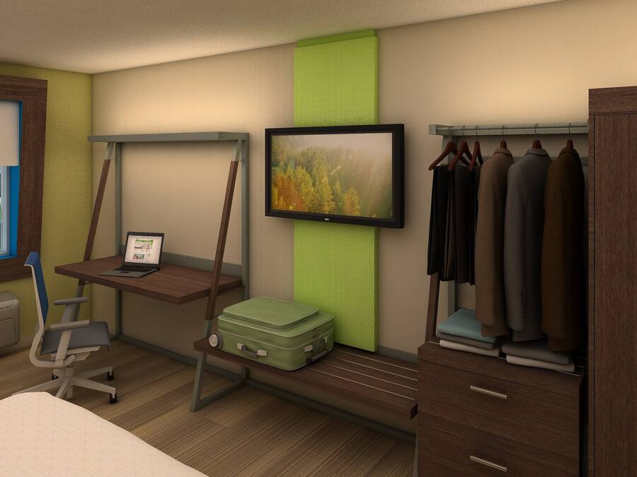 A hotel room with a bed, a desk with a laptop, a green suitcase, a wardrobe, and a TV on the wall.