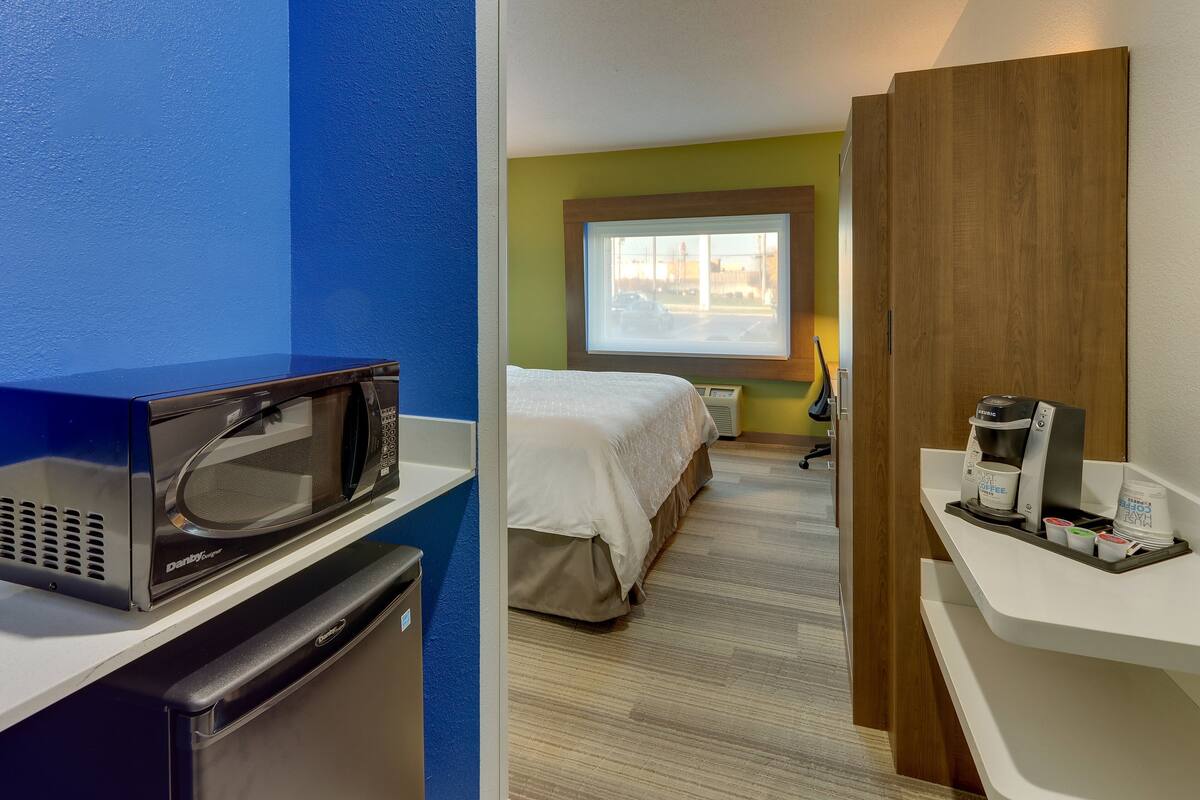 A hotel room with a microwave, a small refrigerator, a bed with a white comforter, a window with a view of the outside, and a coffee machine on a counter.