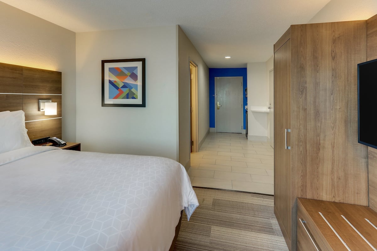 A hotel room with a bed, a nightstand, a wall-mounted lamp, a framed picture, a wooden wardrobe, and a television.