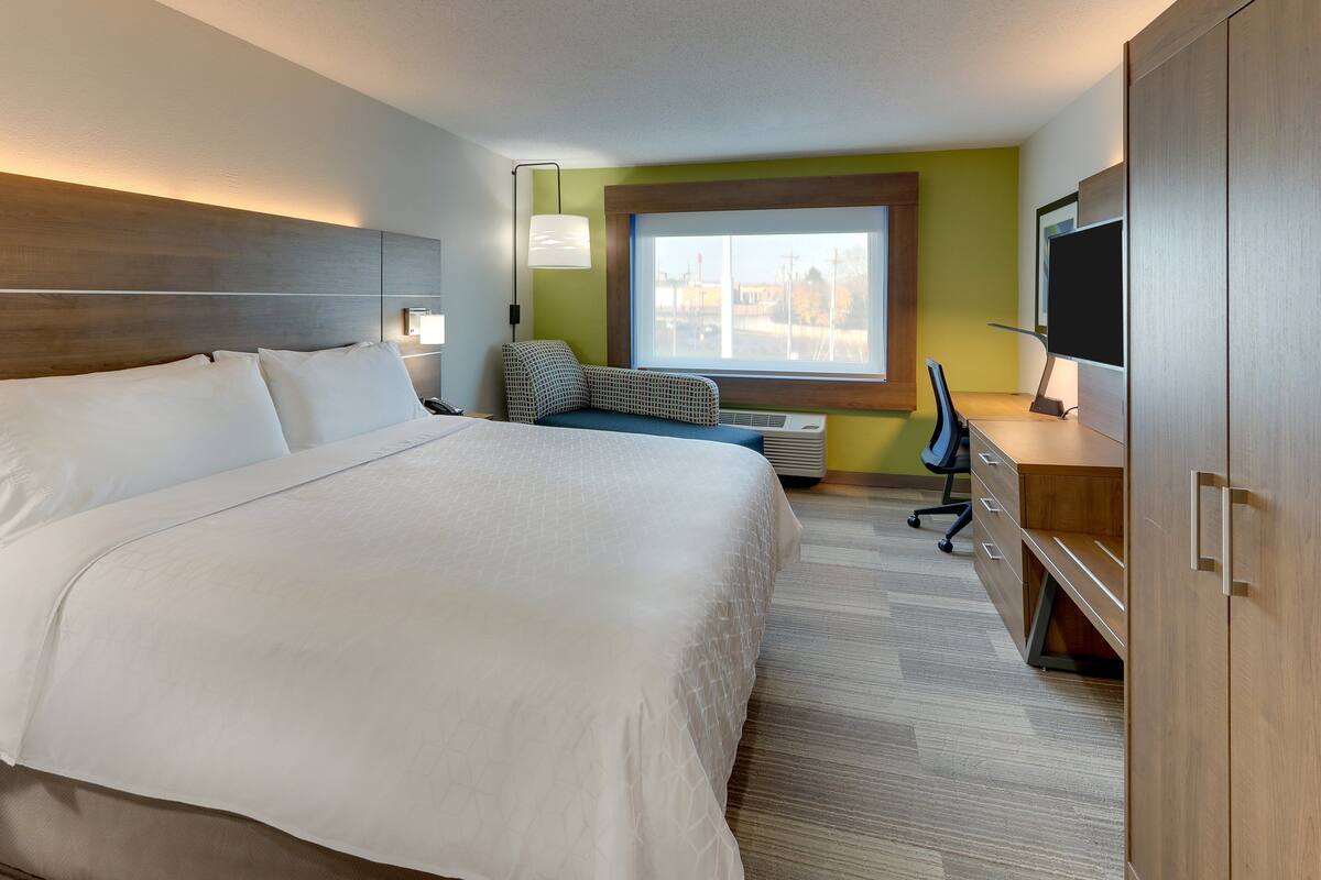 A hotel room with a large bed, a desk with a chair, a TV, and a window with a view of buildings.