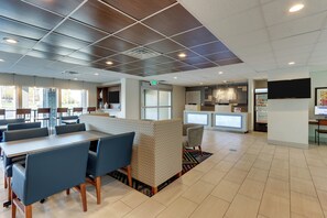 A modern dining area with tables and chairs, a kitchen area with a refrigerator and a flat-screen TV, and a patterned partition. - Holiday Inn Express Cincinnati North - Monroe by IHG (Monroe)