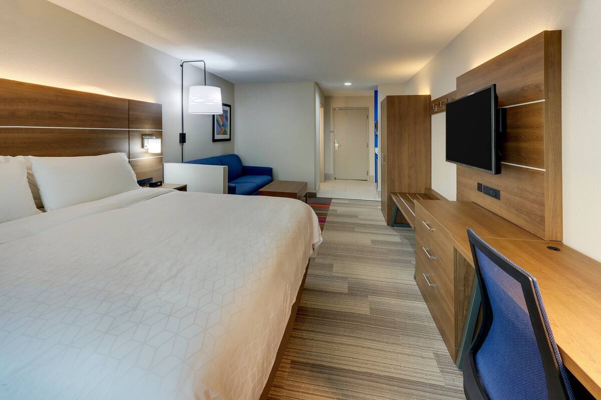 A hotel room with a large bed, a desk, a chair, a television, and a door leading to another room.