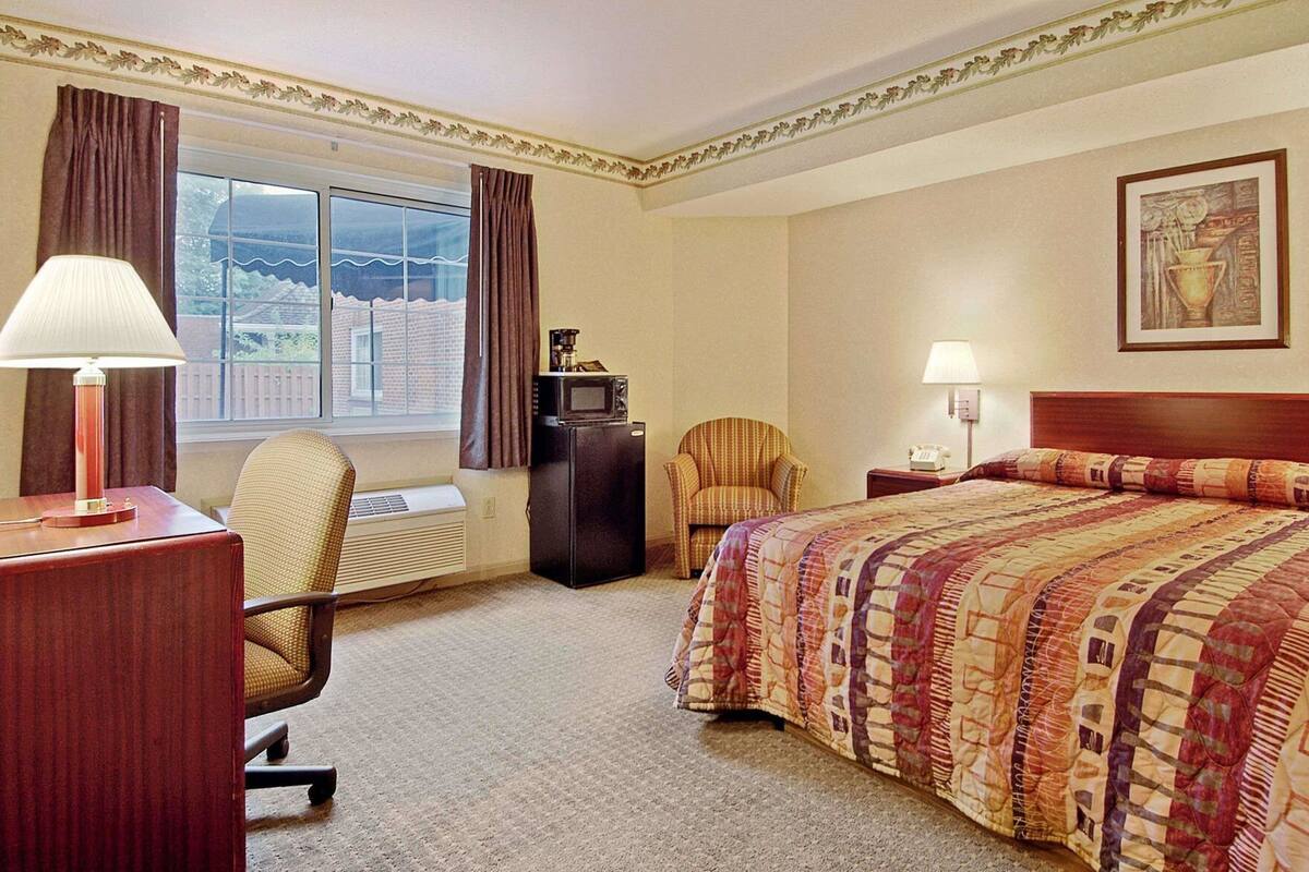 A hotel room with a bed, a desk, a chair, a desk lamp, a lamp, a refrigerator, and a window with curtains.