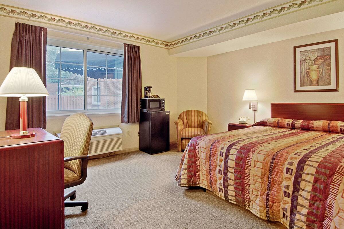 A hotel room with a bed, a desk, a chair, a lamp, a heater, and a window with curtains.