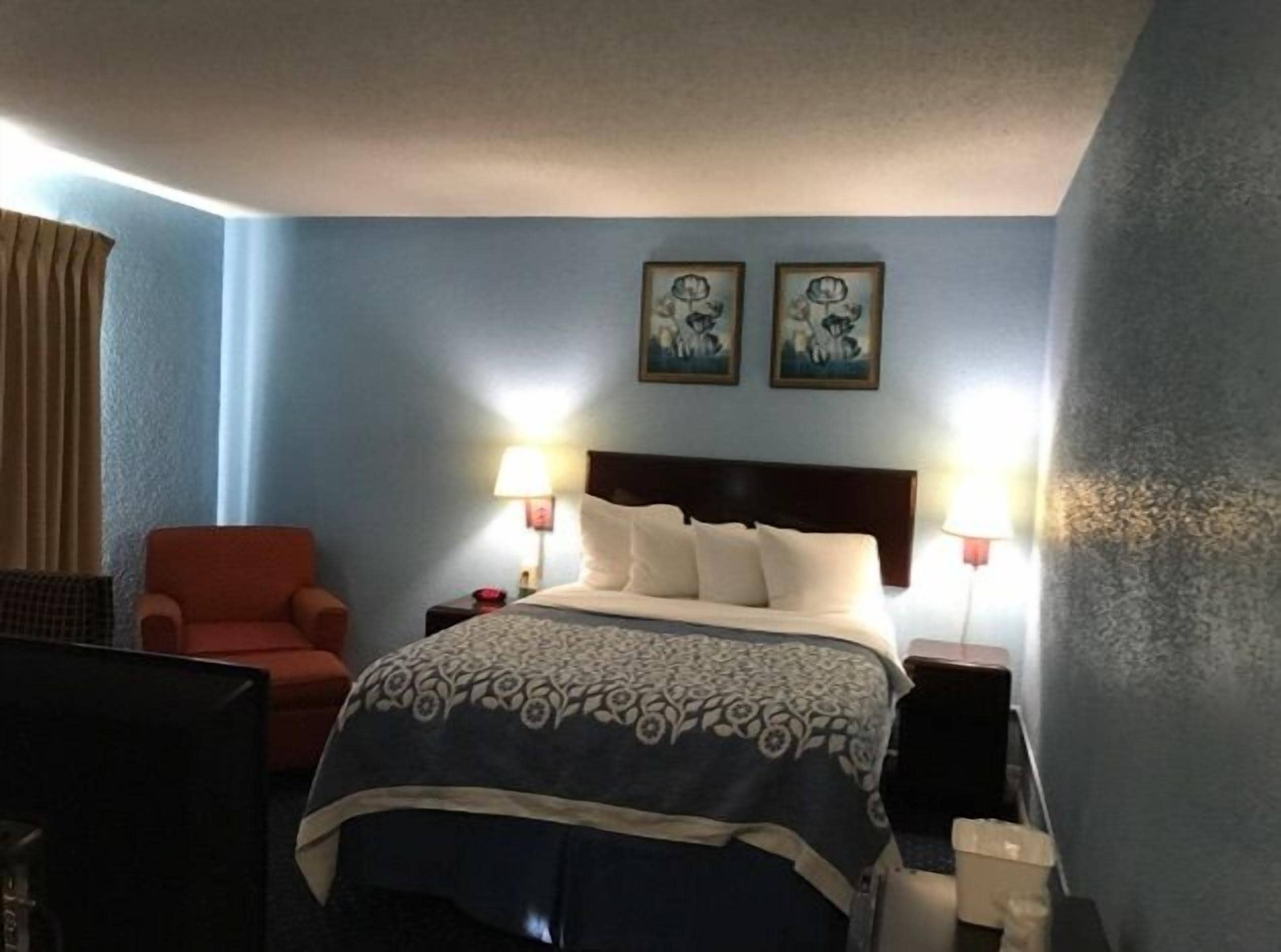 Una habitación de hotel con una cama, una silla, dos lámparas fijadas a la pared y dos cuadros enmarcados.