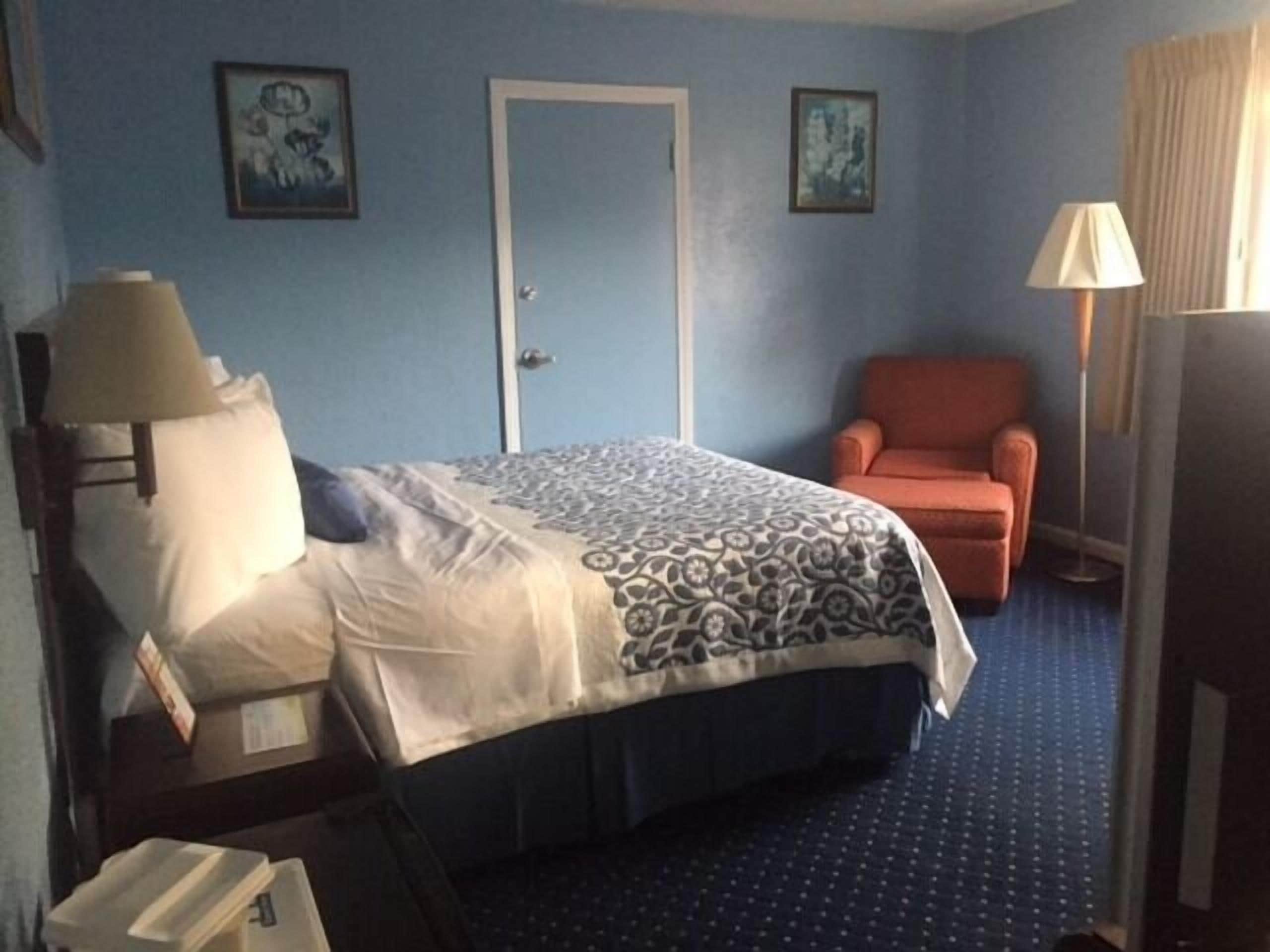 A hotel room with a bed, a chair, a desk with a lamp, and a door.