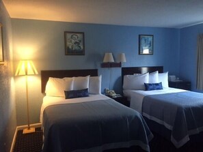 Hotellihuone, jossa on kaksi sänkyä, yöpöytä, yksi lattiavalaisin ja kaksi seinävalaisinta.