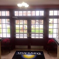 Entrada de hotel con un amplio ventanal, una araña y una alfombra que muestra el logotipo del hotel.
