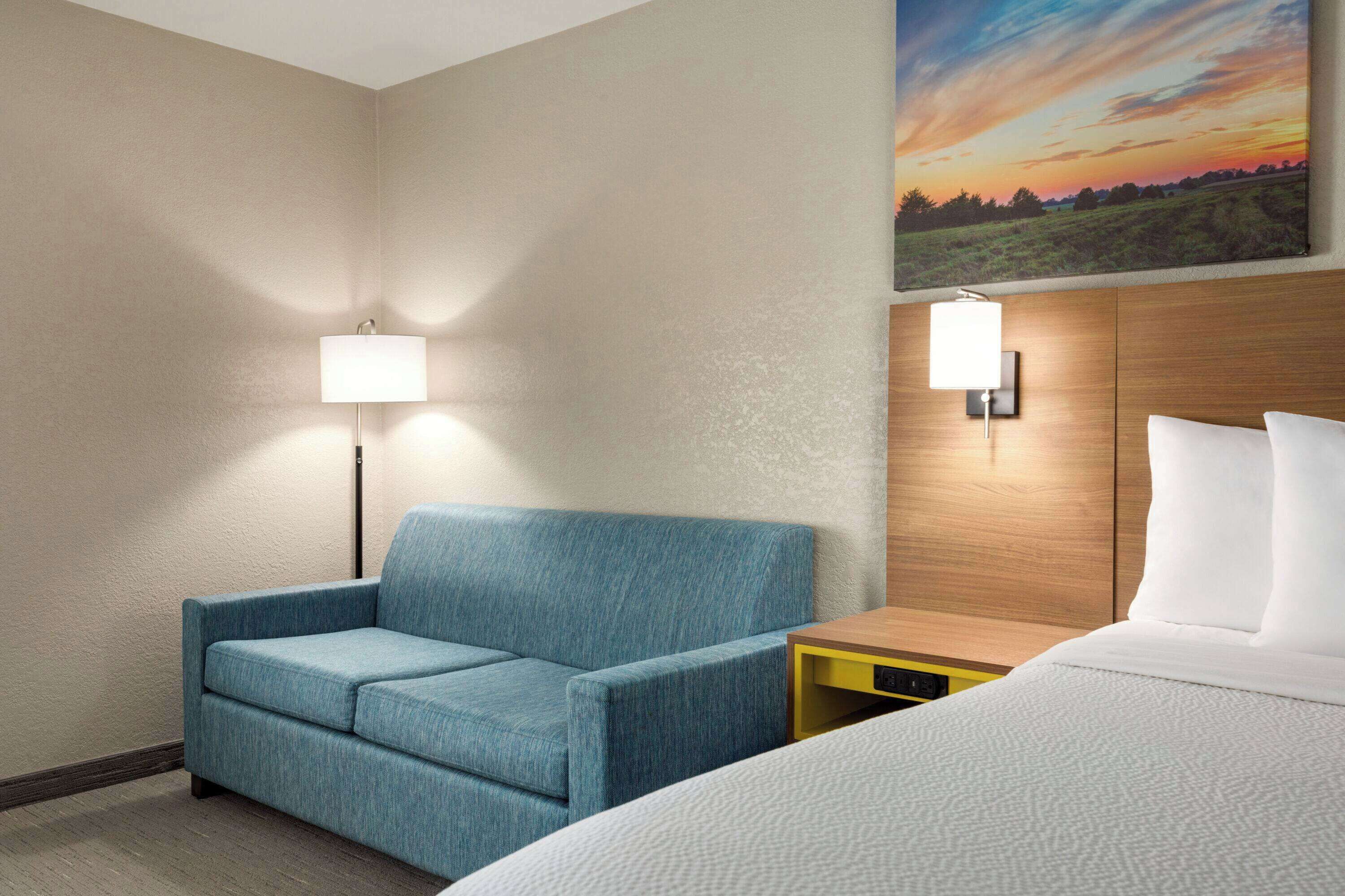 Una habitació d'hotel amb un sofà blau, una tauleta de nit de fusta, un llit amb roba de llit blanca i un quadre amb un paisatge de ponent a la paret.