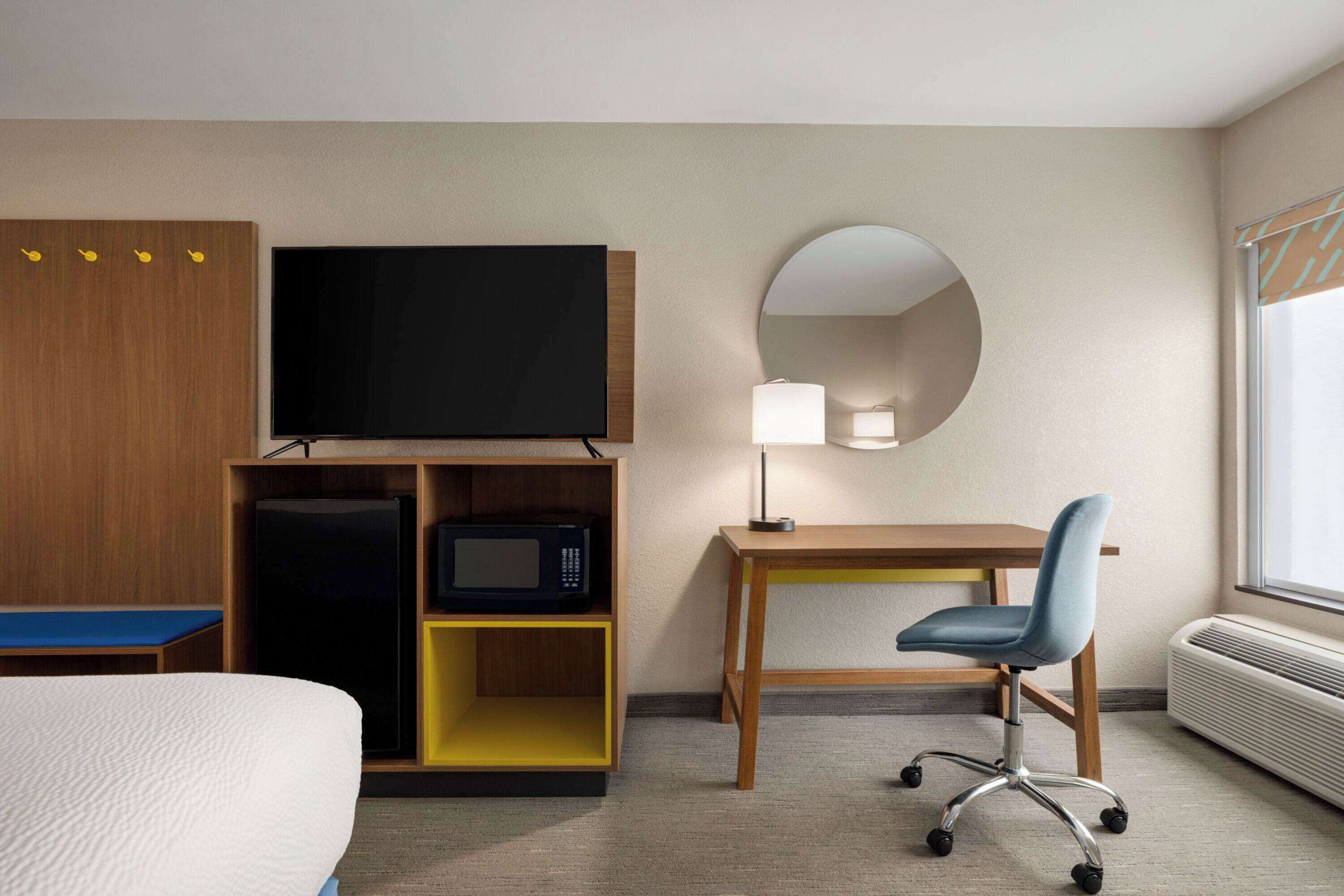 A hotel room with a bed, a desk with a chair, a television, a microwave, a mirror, and a window with a view.
