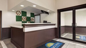 A reception area with a wooden reception desk, a glass door entrance, and a reception desk mat with the logo "Days Inn by Wyndham." - Days Inn by Wyndham Hillsboro (Hillsboro)