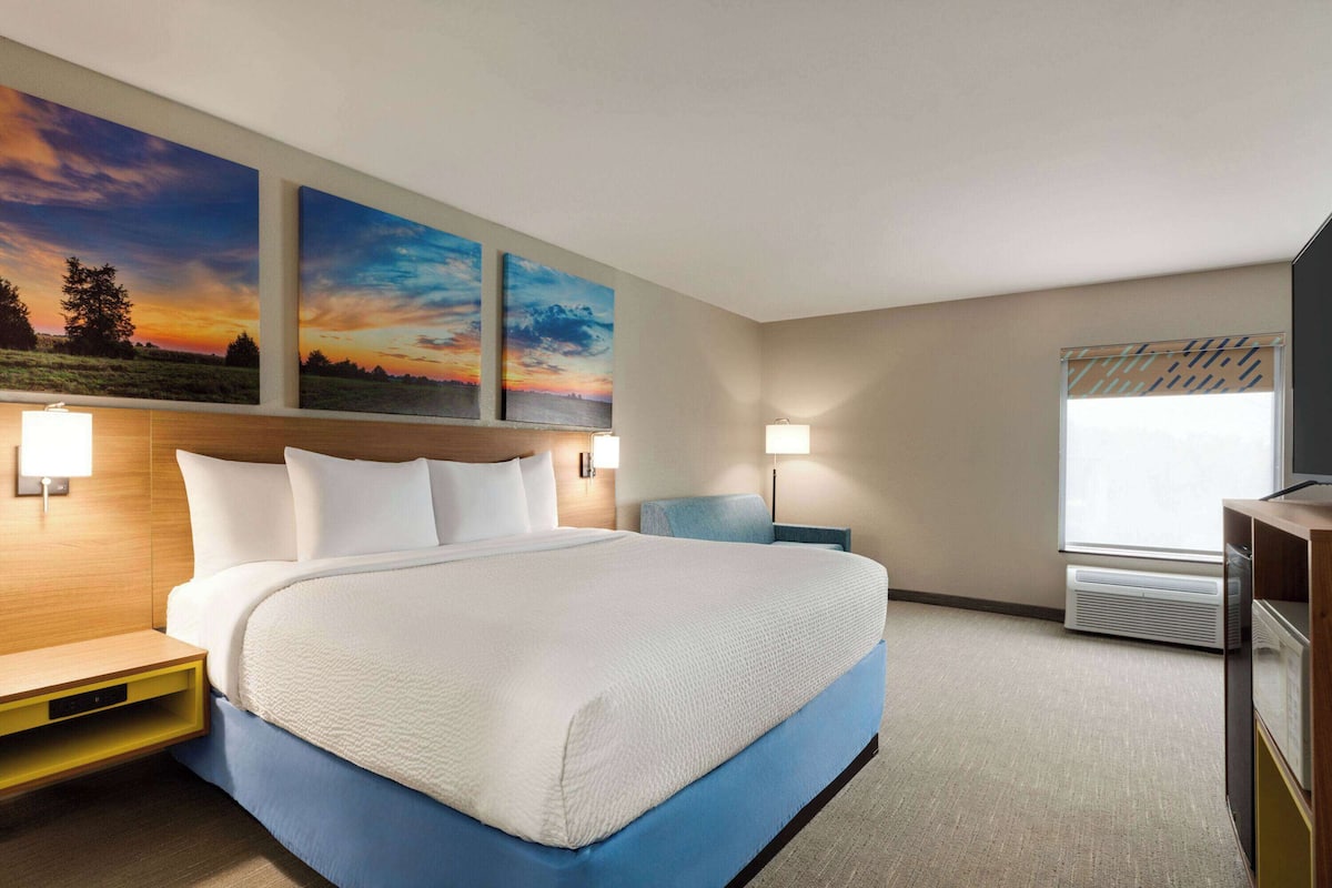 A hotel room with a large bed, two wall-mounted paintings, a nightstand, a desk, and a window with blinds.