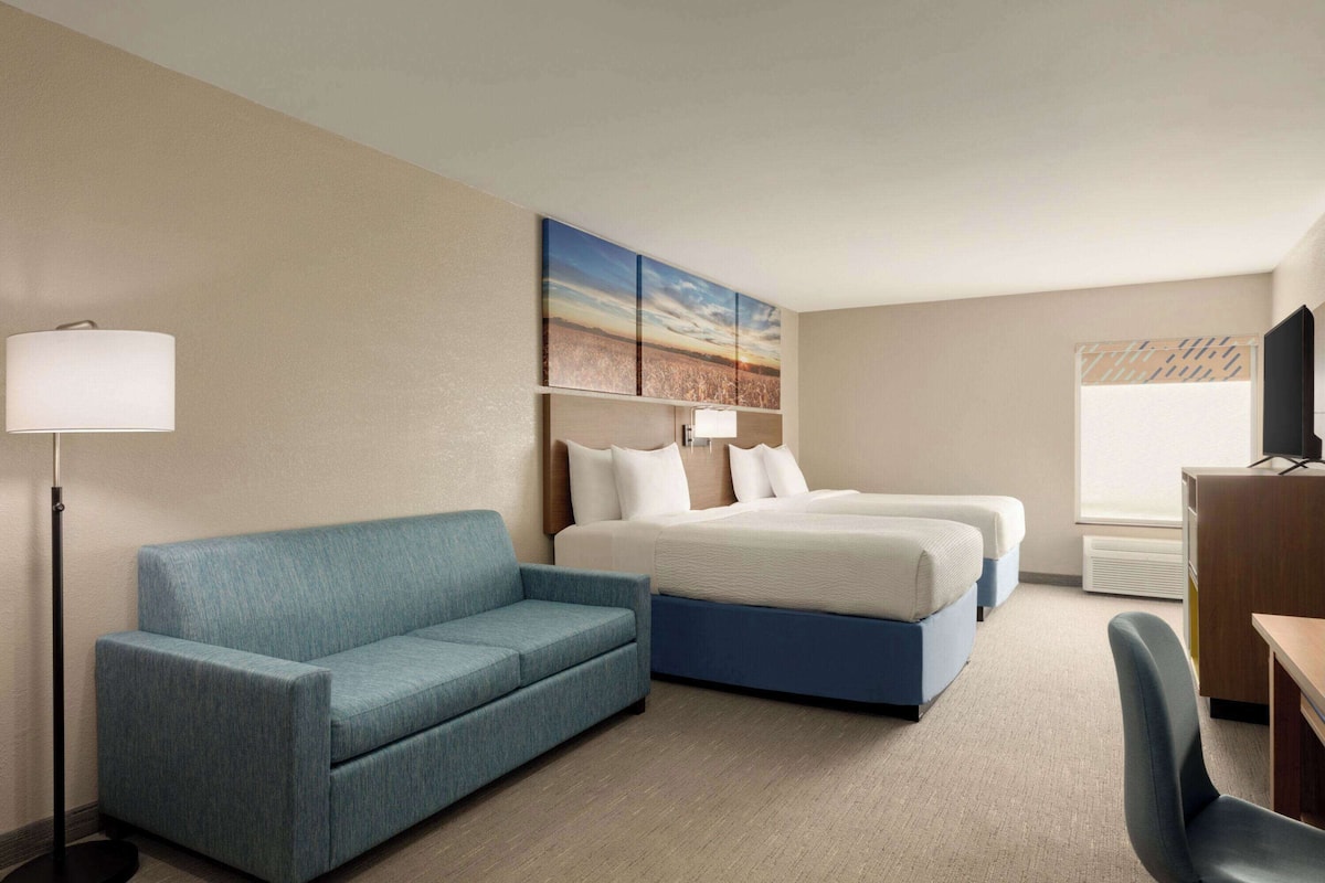 A hotel room with a blue sofa, a bed with white bedding, a desk with a chair, and a painting on the wall.