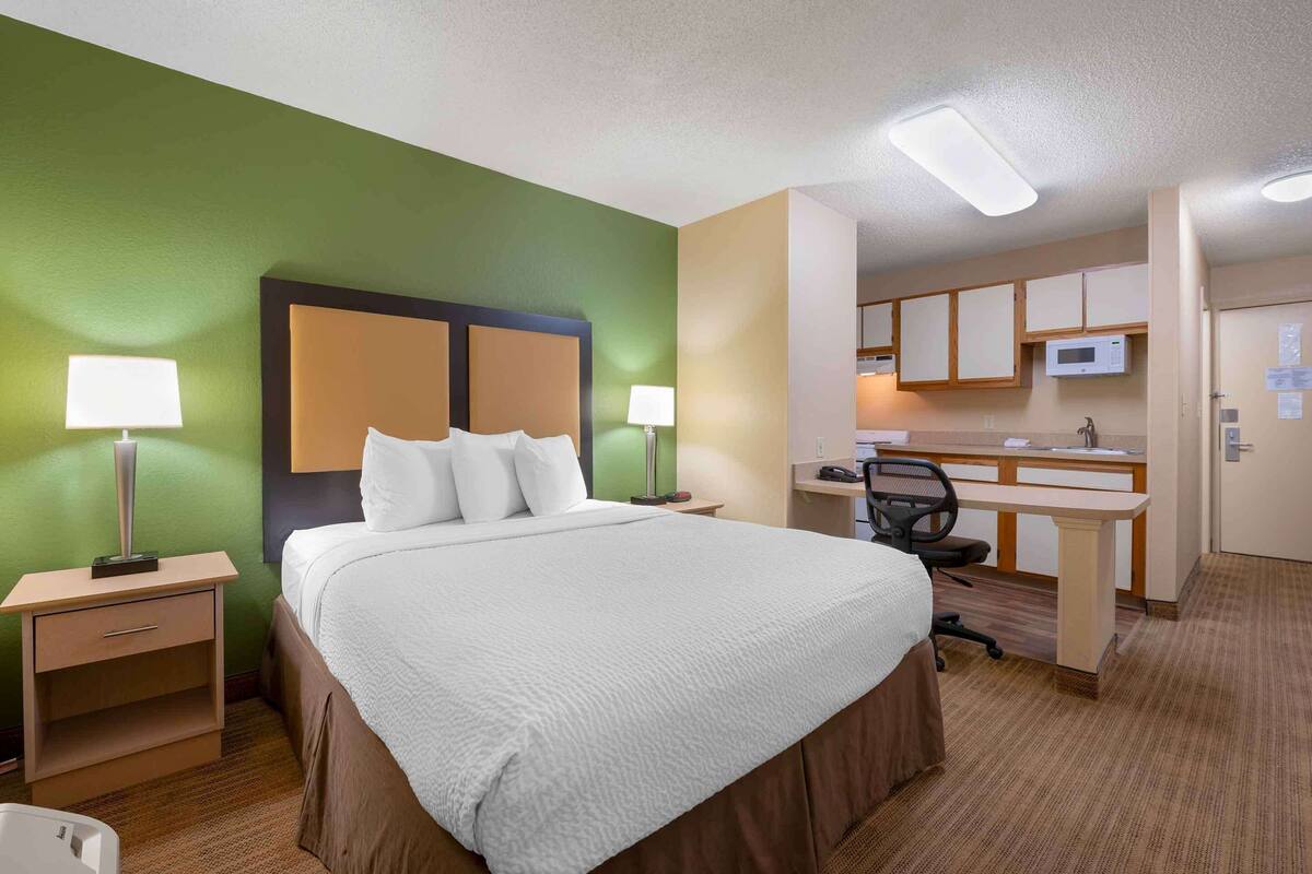 A hotel room with a bed, two bedside tables with lamps, a desk with a chair, a microwave, and a kitchenette.