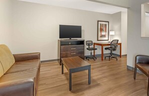A furnished living room with a sofa, a coffee table, a TV, a desk with two chairs, and a framed picture on the wall. - Extended Stay America Suites Dayton Fairborn (Fairborn)