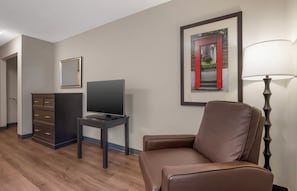 A hotel room with a flat-screen TV, a brown leather chair, a desk with a lamp, a dresser, and framed artwork on the walls.