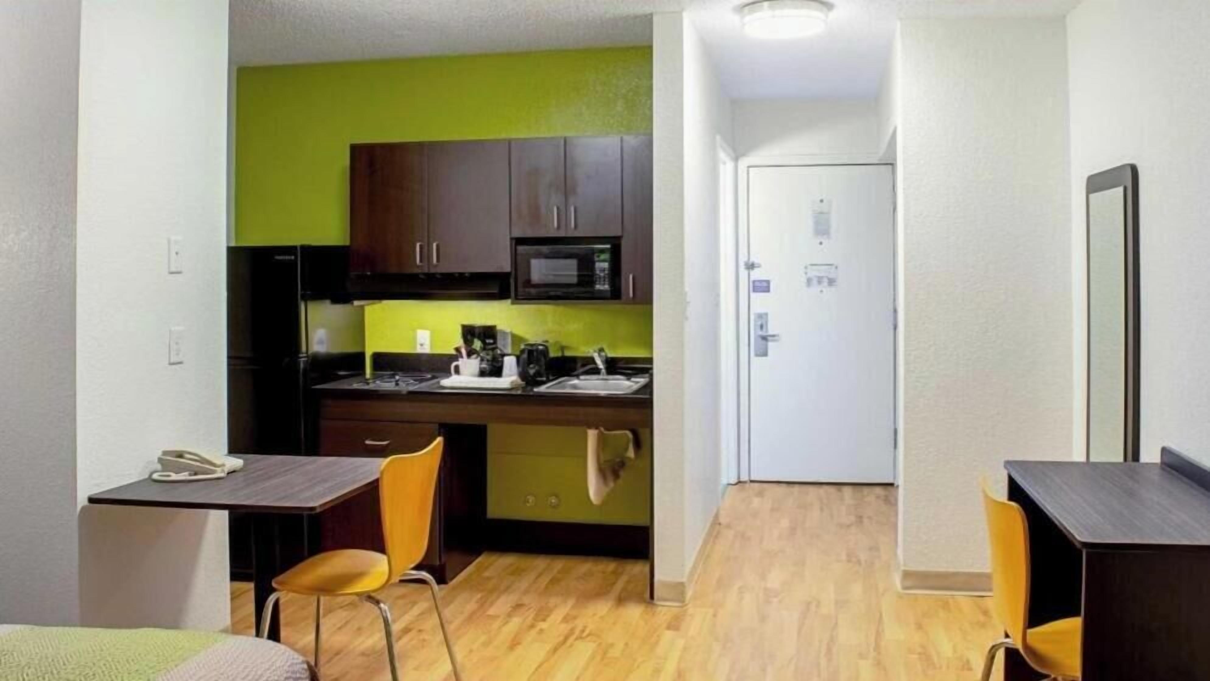 A compact kitchen with a microwave, stove, and sink, a small dining area with chairs, and a door leading to another room.