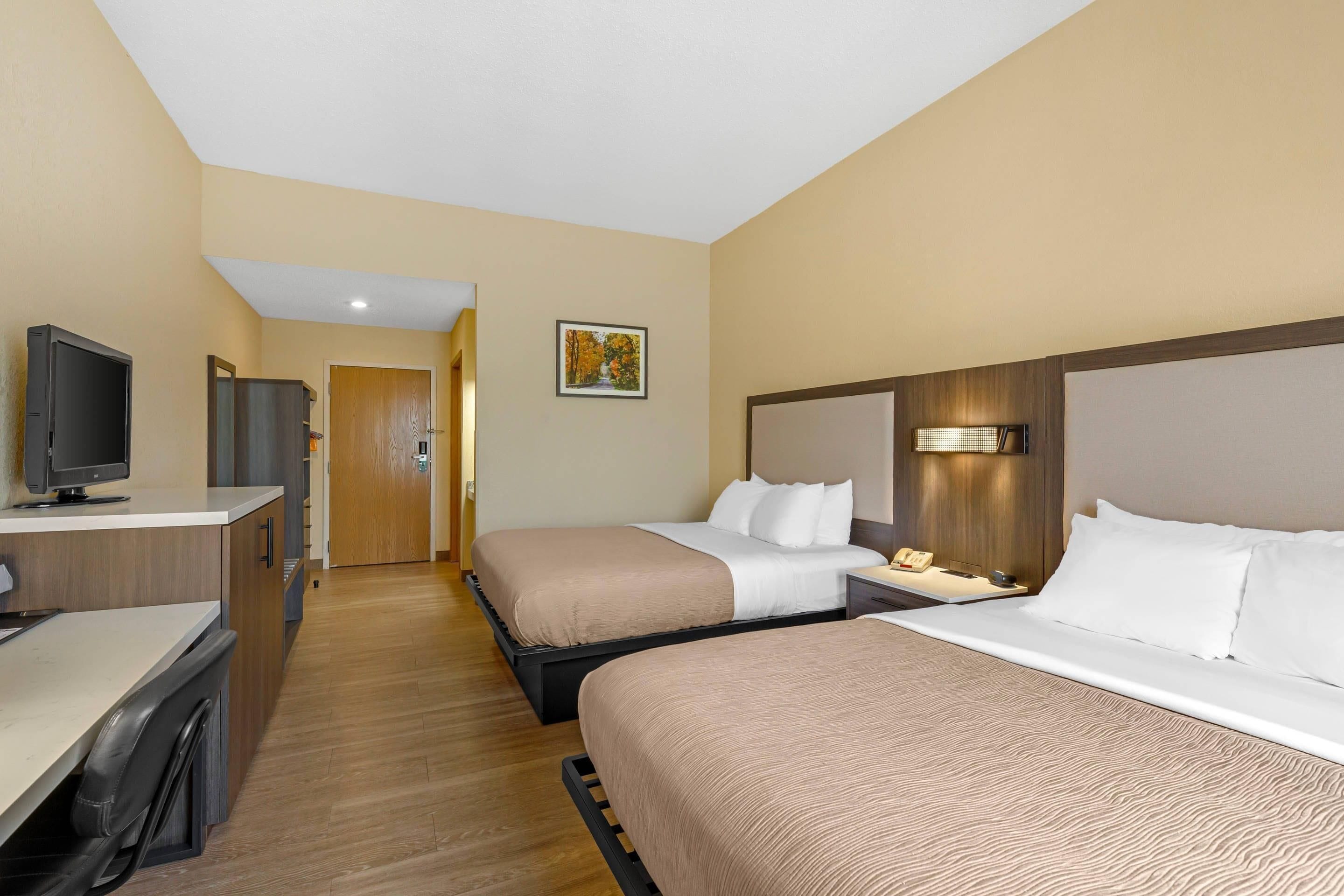 Une chambre d’hôtel avec deux lits, un bureau avec un ordinateur, une télévision et une porte donnant sur une autre pièce.