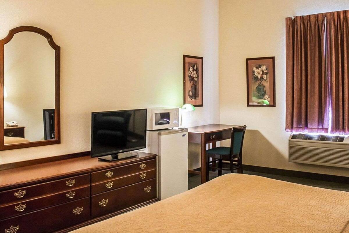 A hotel room with a bed, a desk with a chair, a television, a dresser, a mirror, framed artwork on the wall, and an air conditioning unit.