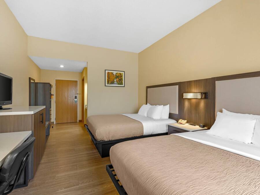 Habitación de hotel con dos camas, un escritorio con una computadora, un televisor y una puerta que da a otra habitación.