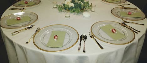 Une table ronde dressée pour un dîner formel, avec des assiettes en verre transparent, des couverts à bord doré et des serviettes vertes.